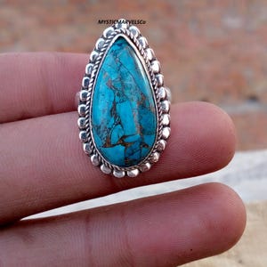 May include: A silver ring with a large, teardrop-shaped turquoise stone with brown veining. The stone is set in a detailed, beaded silver bezel. The ring is a piece of jewellery.