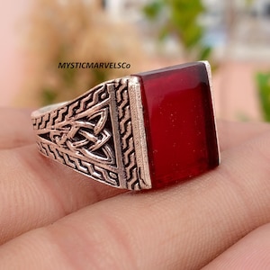 May include: A silver-toned ring featuring a large, rectangular, deep red gemstone set into a band with intricate, raised, dark-oxidized metal patterns and a Celtic-inspired knot design on the side. The text MYSTICMARVELSCO is visible.