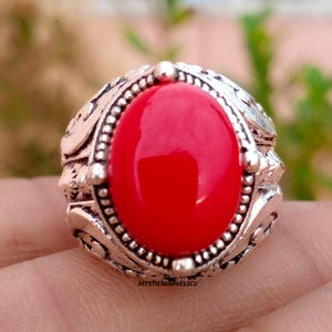 May include: A silver-toned ring featuring a large, smooth, oval-shaped red stone set in the center. The metal band displays intricate, raised scrollwork patterns and a beaded border surrounding the stone. The text MYSTICMARVELSCO is visible.