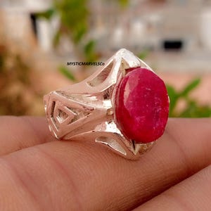 May include: A silver ring featuring a large, oval-shaped, deep red gemstone set in a band with geometric cut-out patterns. The text MYSTICMARVELSCO is visible on the side of the metal band. The ring has a polished finish and a bold design.