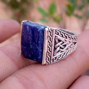 May include: A silver-toned ring featuring a rectangular, deep blue lapis lazuli stone set in the center. The band is crafted with intricate, raised metalwork patterns, including a prominent triangular knot design on the side of the shank.