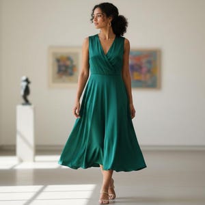 May include: A sleeveless, emerald green dress with a V-neck and a flowing skirt. The dress is knee-length and worn by a person walking in a gallery. The dress has a wrap-style bodice and a subtle sheen.
