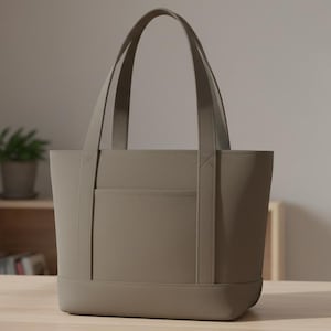 May include: A taupe tote bag with two handles and a front pocket. The bag is made of a smooth material and sits on a light-colored wooden surface. The background includes a small plant in a pot and a wooden shelf.