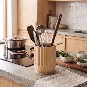 May include: A cylindrical, ribbed, beige kitchen utensil holder containing a dark wood spatula, a metal ladle, metal tongs, a wire whisk, and a long dark wood spoon. The holder has a smooth, rounded base and vertical grooves along its exterior surface.