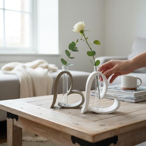 May include: Two heart-shaped vases, one grey and one white, each holding a single stem flower. The vases sit on a wooden table, with a white rose and greenery in one. A hand reaches towards the white vase.