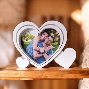 May include: A white heart-shaped photo frame with a picture of a couple. The frame has a layered design and two small heart accents. The photo shows a man and woman smiling. The frame is on a wooden surface.