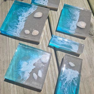 Könnte beinhalten: Ein Set aus fünf Untersetzern und Haltern aus Kunstharz im Strand-Design mit durchscheinendem türkisfarbenem Wasser, weißen Schaumwellen und eingebetteten Muscheln auf einem sandbeigen Untergrund in verschiedenen Formen.