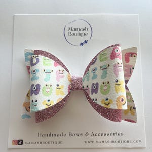 May include: A hair bow featuring a white base with a colourful pattern of small cartoon monsters and letters, layered with pink glitter fabric. The bow is attached to a display card labelled Mamash Boutique Handmade Bows & Accessories.