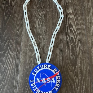 May include: A white plastic chain necklace with a blue circular pendant. The pendant features the NASA logo, the words "FUTURE ROCKET SCIENTIST" in white, and a red arrow. The necklace is displayed on a wooden surface.