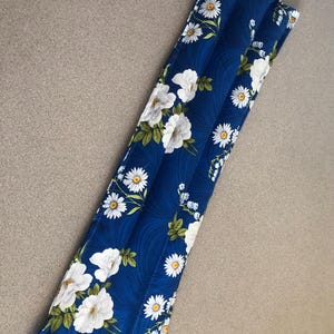 May include: A long, rectangular, blue heat pack with a floral pattern of white daisies and roses. The fabric has a wavy design. The heat pack is filled with a substance to provide warmth and comfort.