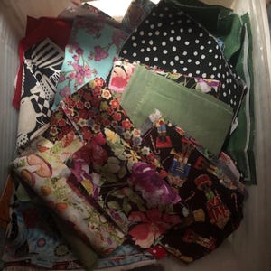 May include: A collection of assorted fabric scraps in a white plastic bin. The pieces feature various patterns including black and white polka dots, floral prints in red, pink, and blue, mushroom illustrations, and nutcracker figures on black.