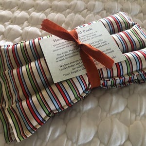 May include: A striped heat and cold pack tied with an orange ribbon. The pack features vertical stripes in red, blue, green, and beige. A small tag with instructions is attached to the pack.