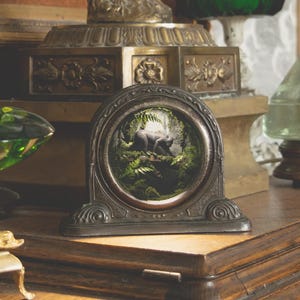 Puede incluir: Una mesa de madera exhibe objetos antiguos. Incluye un cuenco de vidrio verde, una pequeña imagen enmarcada, un candelabro de latón, una estatua dorada, un espejo pequeño, una lámpara de vidrio verde y un jarrón blanco con flores amarillas.