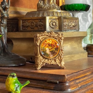 Pode incluir: Uma coleção de objetos decorativos incluindo um candeeiro figurativo de latão, um pequeno relógio dourado ornamentado com um mostrador circular de paisagem, uma taça de vidro verde, uma pirâmide de vidro verde, um pássaro verde e um espelho oval.
