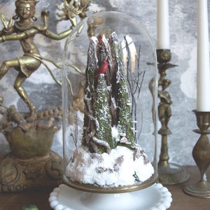 Op de afbeelding: Een decoratieve glazen stolp met een miniatuur winters tafereel. Binnenin toont een besneeuwd landschap miniatuurbomen en twee rode kardinalen. De stolp staat op een witte, met kralen versierde basis. Twee witte kaarsen in messing houders staan op de achtergrond.