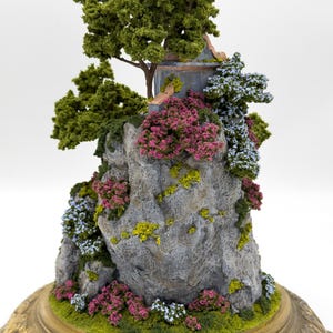 May include: A miniature scene of a small stone cliff with a crumbling building on top. The cliff is covered in green moss and small pink and blue flowers. A small green tree grows on the cliff. The scene is set on a gold-colored base.