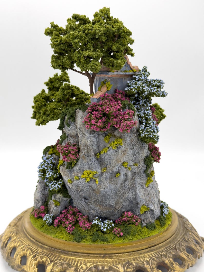 May include: A miniature scene of a small stone cliff with a crumbling building on top. The cliff is covered in green moss and small pink and blue flowers. A small green tree grows on the cliff. The scene is set on a gold-colored base.