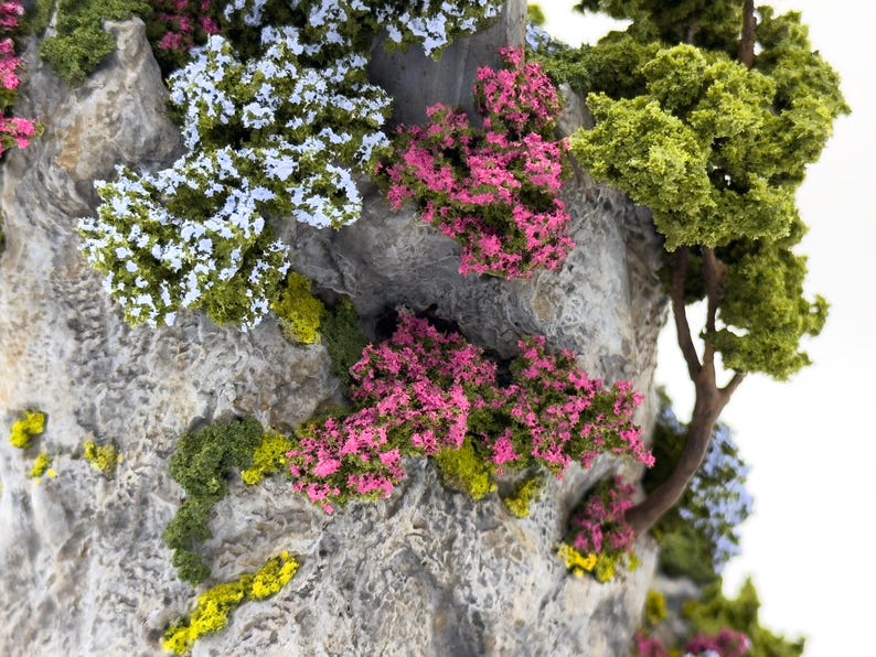May include: A close-up of a miniature rock formation with a variety of green, pink, and white foliage. The rock is textured and gray.