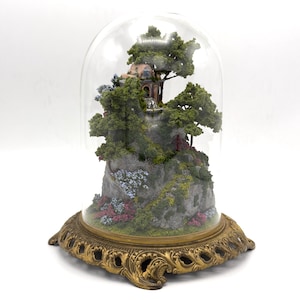 May include: A miniature glass dome with a gold base, containing a small, detailed model of a house on a rocky hill with trees and flowers.