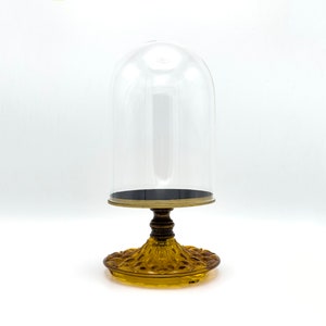 May include: A clear glass dome sits atop a decorative amber glass base. The dome is designed to protect and display small items.