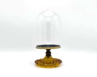 May include: A clear glass dome sits atop a decorative amber glass base. The dome is designed to protect and display small items.