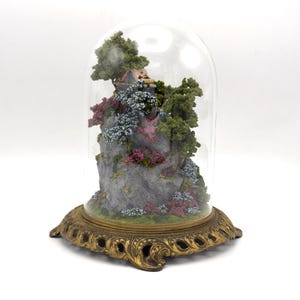 May include: A miniature glass dome with a gold metal base, containing a small house on a rocky cliff with trees and flowers.