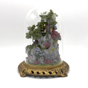 May include: A miniature landscape scene with a small house, trees, and flowers, encased in a glass dome on a gold metal base.