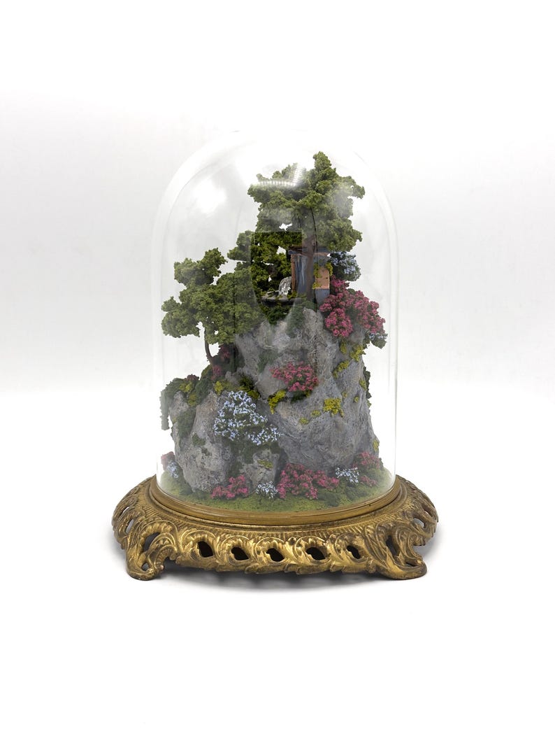 May include: A miniature landscape scene with a small house, trees, and flowers, encased in a glass dome on a gold metal base.