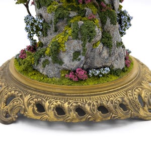 May include: A miniature rock formation with a small tree growing out of the top. The rock is covered in moss and small flowers. The base is a gold metal stand with a decorative design.
