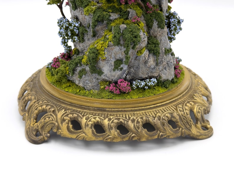 May include: A miniature rock formation with a small tree growing out of the top. The rock is covered in moss and small flowers. The base is a gold metal stand with a decorative design.