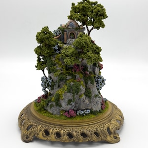 May include: A miniature stone house with a blue door and a brown roof sits on a rocky cliff covered in green moss and small pink and blue flowers. The cliff is on a gold metal base with a decorative design.