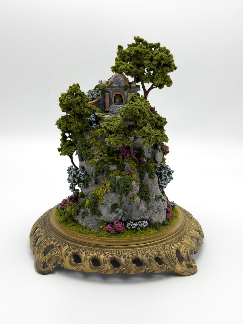 May include: A miniature stone house with a blue door and a brown roof sits on a rocky cliff covered in green moss and small pink and blue flowers. The cliff is on a gold metal base with a decorative design.