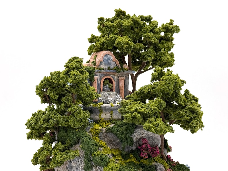 May include: A miniature stone building with a fountain and a domed roof, surrounded by lush green trees and foliage. The building is decorated with red and brown accents, and the fountain is flowing with water.