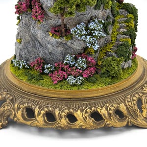 May include: A miniature landscape scene with a rocky mountain, green foliage, and pink and blue flowers. The scene is displayed on a gold-colored, ornate base.