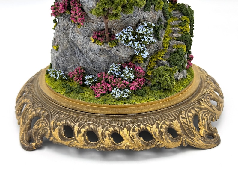 May include: A miniature landscape scene with a rocky mountain, green foliage, and pink and blue flowers. The scene is displayed on a gold-colored, ornate base.