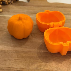 May include: Two bright orange pumpkin-shaped containers. One container is closed, showing a ribbed exterior with a small stem on top. The second container is open, revealing a hollow interior with a hinged lid and a smooth, rounded base structure.