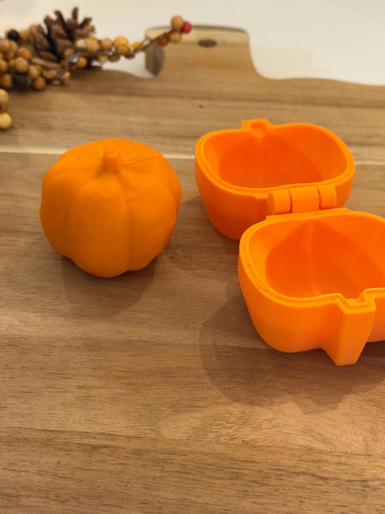May include: Two bright orange pumpkin-shaped containers. One container is closed, showing a ribbed exterior with a small stem on top. The second container is open, revealing a hollow interior with a hinged lid and a smooth, rounded base structure.
