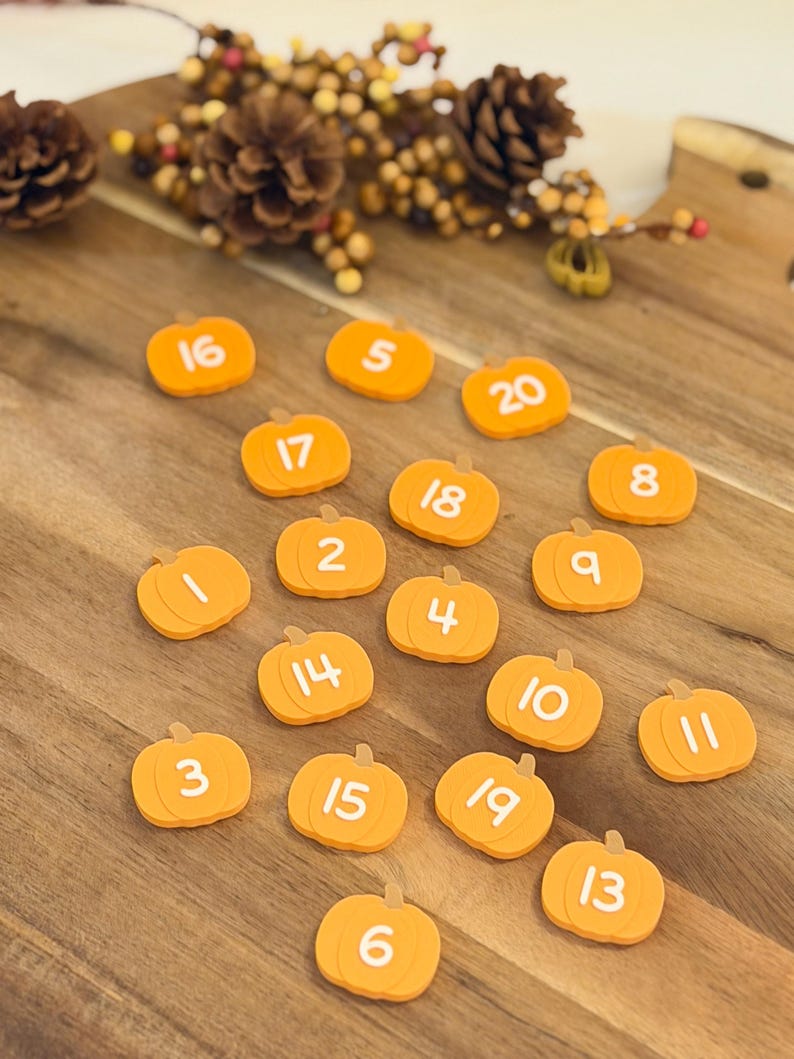 May include: A collection of twenty small, orange, pumpkin-shaped tokens, each featuring a unique white number from 1 to 20 printed on the front. The pumpkin tokens are arranged in a scattered pattern, showcasing a smooth, flat surface design.
