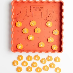 May include: A square orange tray with a scalloped edge featuring a raised pumpkin patch design and the text PUMPKIN PATCH. The tray includes alphabet letters embossed on the surface and a set of loose, small orange pumpkin-shaped letter tokens.