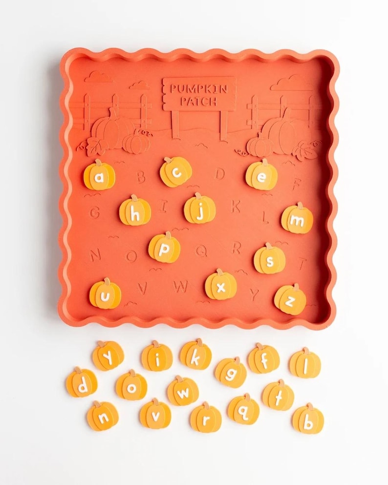 May include: A square orange tray with a scalloped edge featuring a raised pumpkin patch design and the text PUMPKIN PATCH. The tray includes alphabet letters embossed on the surface and a set of loose, small orange pumpkin-shaped letter tokens.