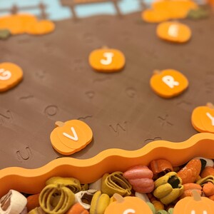 May include: A brown rectangular tray with a wavy orange border featuring a pumpkin patch scene with embossed letters on the surface. Small orange pumpkin-shaped tokens with white letters are scattered across the tray and in a pile below.