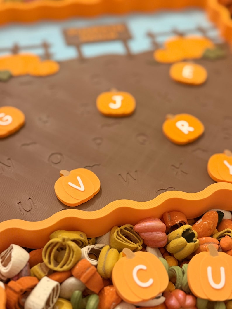 May include: A brown rectangular tray with a wavy orange border featuring a pumpkin patch scene with embossed letters on the surface. Small orange pumpkin-shaped tokens with white letters are scattered across the tray and in a pile below.