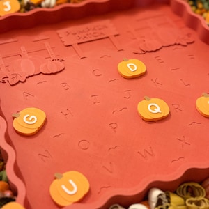 May include: A square, terracotta-colored tray with a scalloped edge featuring an embossed pumpkin patch scene and the alphabet. The tray includes loose, orange pumpkin-shaped tokens marked with white letters, including D, G, Q, U, and Y.