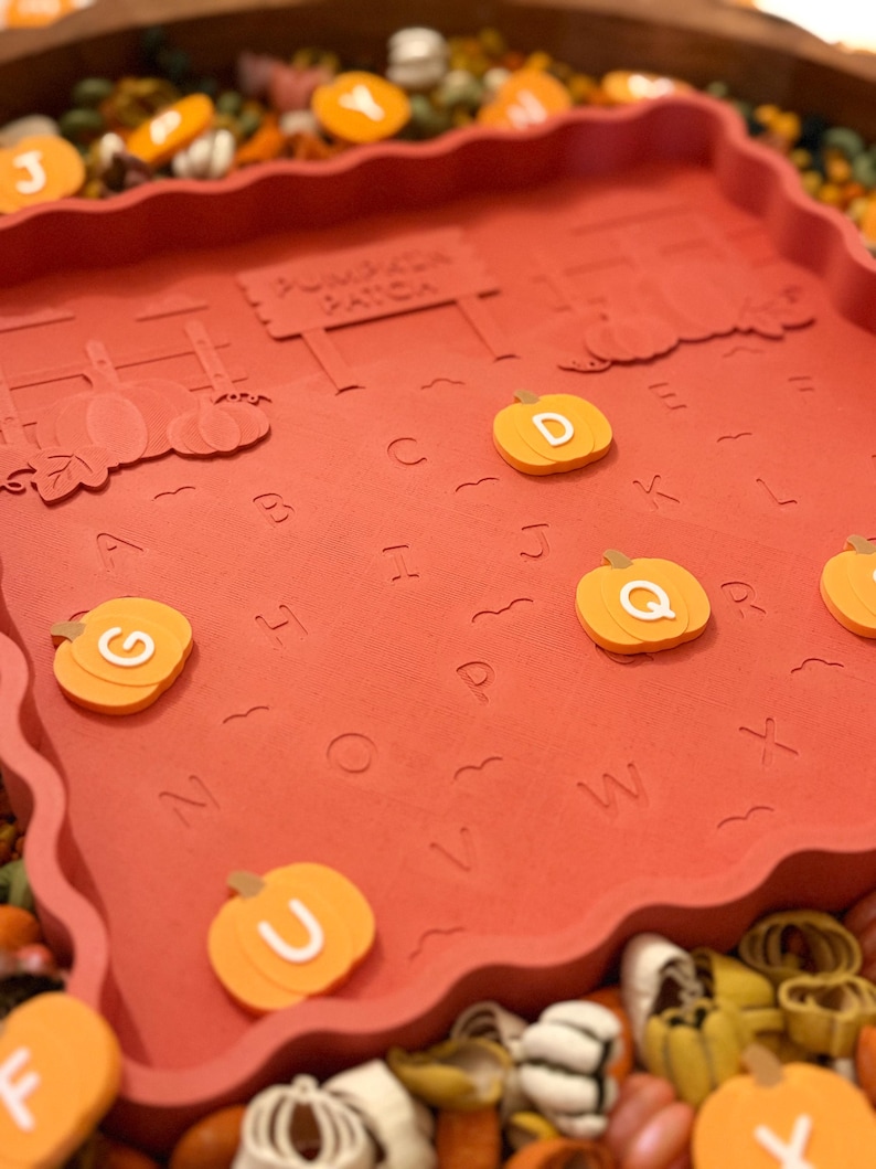May include: A square, terracotta-colored tray with a scalloped edge featuring an embossed pumpkin patch scene and the alphabet. The tray includes loose, orange pumpkin-shaped tokens marked with white letters, including D, G, Q, U, and Y.