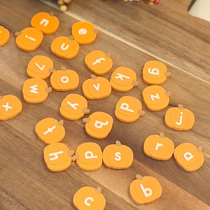 May include: A collection of small, orange, pumpkin-shaped tokens, each featuring a single white lowercase letter printed on its surface. These individual pumpkin pieces are scattered across a wooden surface, displaying various letters of the alphabet.