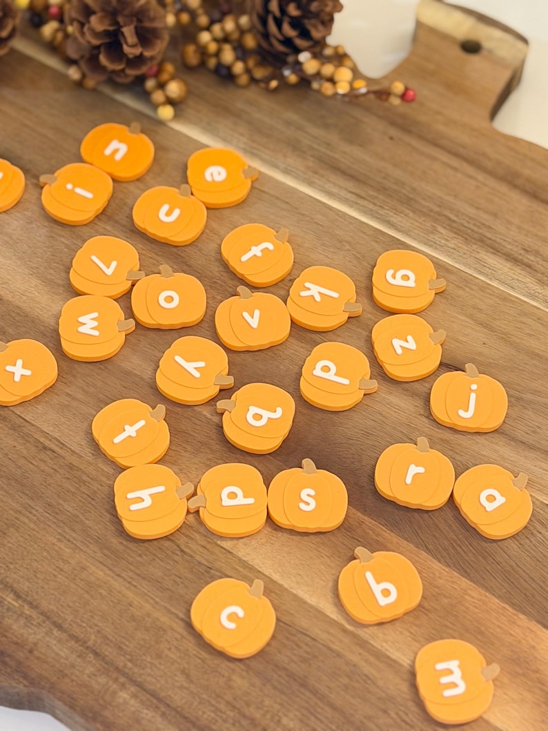 May include: A collection of small, orange, pumpkin-shaped tokens, each featuring a single white lowercase letter printed on its surface. These individual pumpkin pieces are scattered across a wooden surface, displaying various letters of the alphabet.
