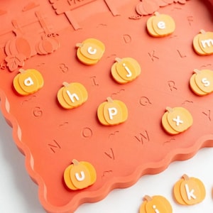 May include: A rectangular orange tray with a scalloped edge featuring a raised PUMPKIN PATCH sign and embossed letters. Several orange pumpkin-shaped pieces with white lowercase letters are placed on the tray, with additional pieces nearby.