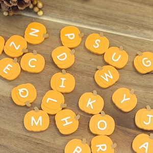 May include: A collection of small, orange, pumpkin-shaped flat ornaments, each featuring a single white capital letter printed on the front. The pumpkins are arranged in a scattered pattern, displaying various letters of the alphabet across the surface.