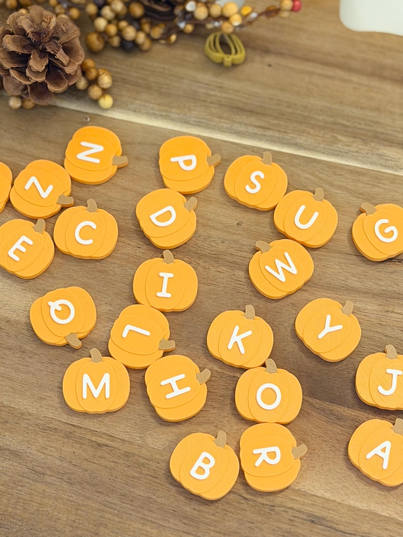 May include: A collection of small, orange, pumpkin-shaped flat ornaments, each featuring a single white capital letter printed on the front. The pumpkins are arranged in a scattered pattern, displaying various letters of the alphabet across the surface.