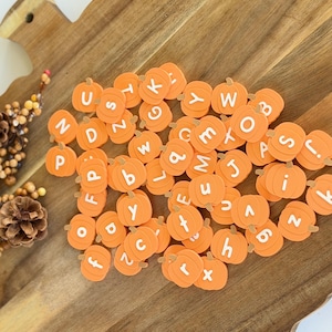 May include: A collection of small, orange, pumpkin-shaped beads, each featuring a single white letter printed on the front. The pumpkin beads are scattered across a wooden surface, displaying a variety of uppercase and lowercase alphabet letters.